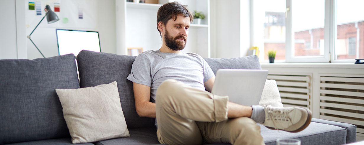 Young relaxed man sitting on sofa with laptop in front and watching webcast or scrolling in the net