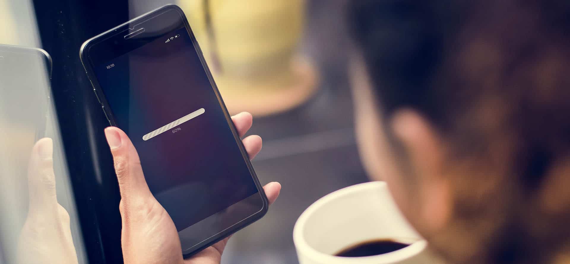 A person looks at a smartphone screen displaying a loading bar, holding a coffee cup in the other hand, with a blurred background emphasizing focus on the phone.