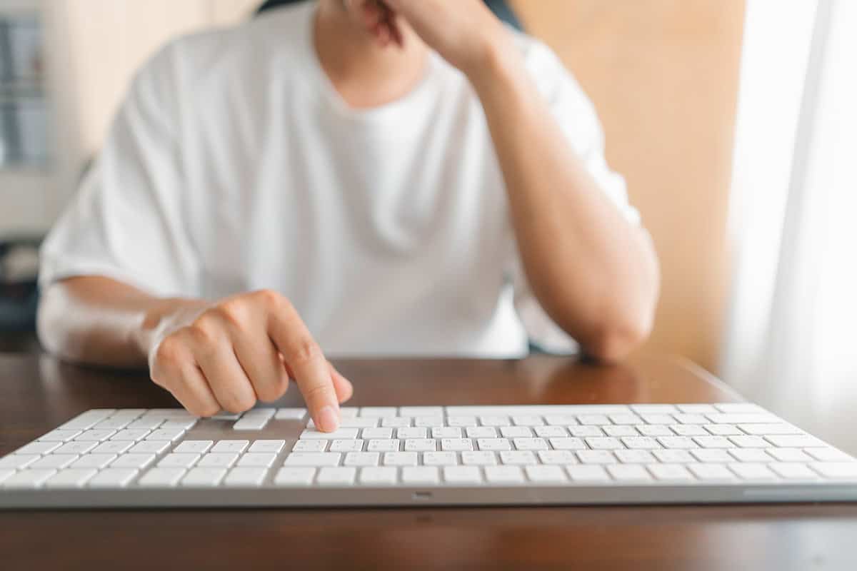A person in a white shirt is typing on a white keyboard, with only their hands and lower face visible, sitting at a wooden desk.