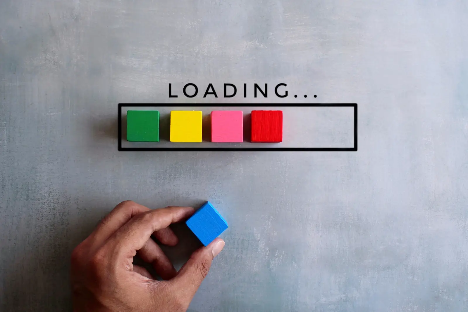 A person's hand placing a blue cube alongside other colorful cubes arranged to resemble a loading bar that spells out "loading..." against a gray background.