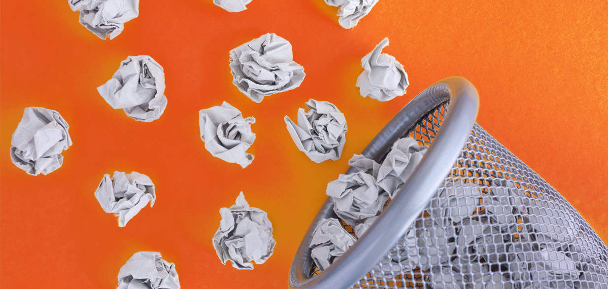Crumpled white paper balls being tossed into a silver mesh wastebasket, with some missing and scattered on a vibrant orange background.